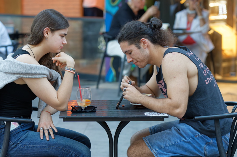 20 - A young woman with long dark hair and a young man with a man bun sitting at an outdoor cafe table, with drinks and a smartphone on the table